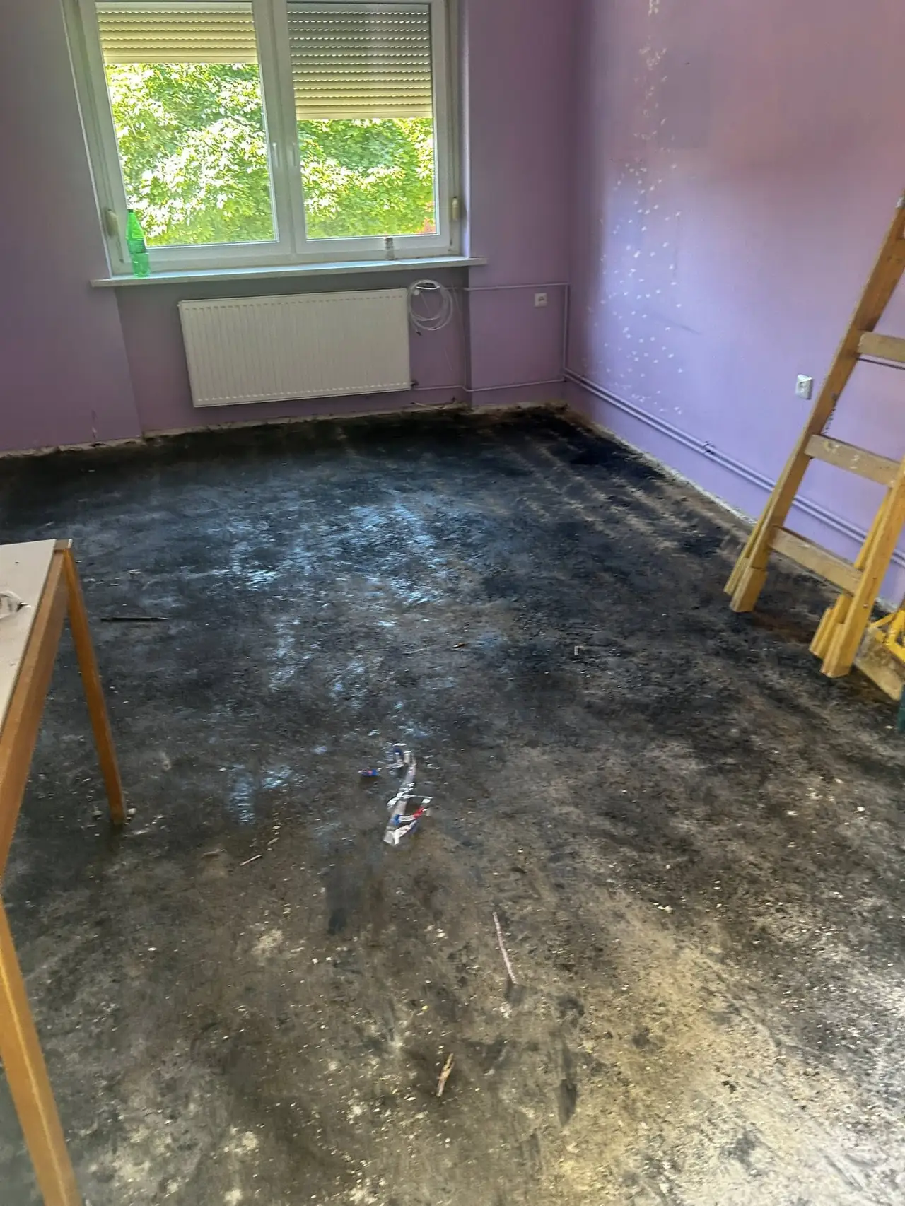 Üres, koszos padlójú helyiség látható, amelynek falai világos lila színűek. Az ablakon át természetes fény áramlik be, és a szoba egyik sarkában egy létra áll. A padló felújítása folyamatban van, amit a Farkas-Konstrukt Kft. generálkivitelezési szolgáltatásaival végeznek Győrben. A környezet rendezetlen, de a felújítás jelei már látszanak.