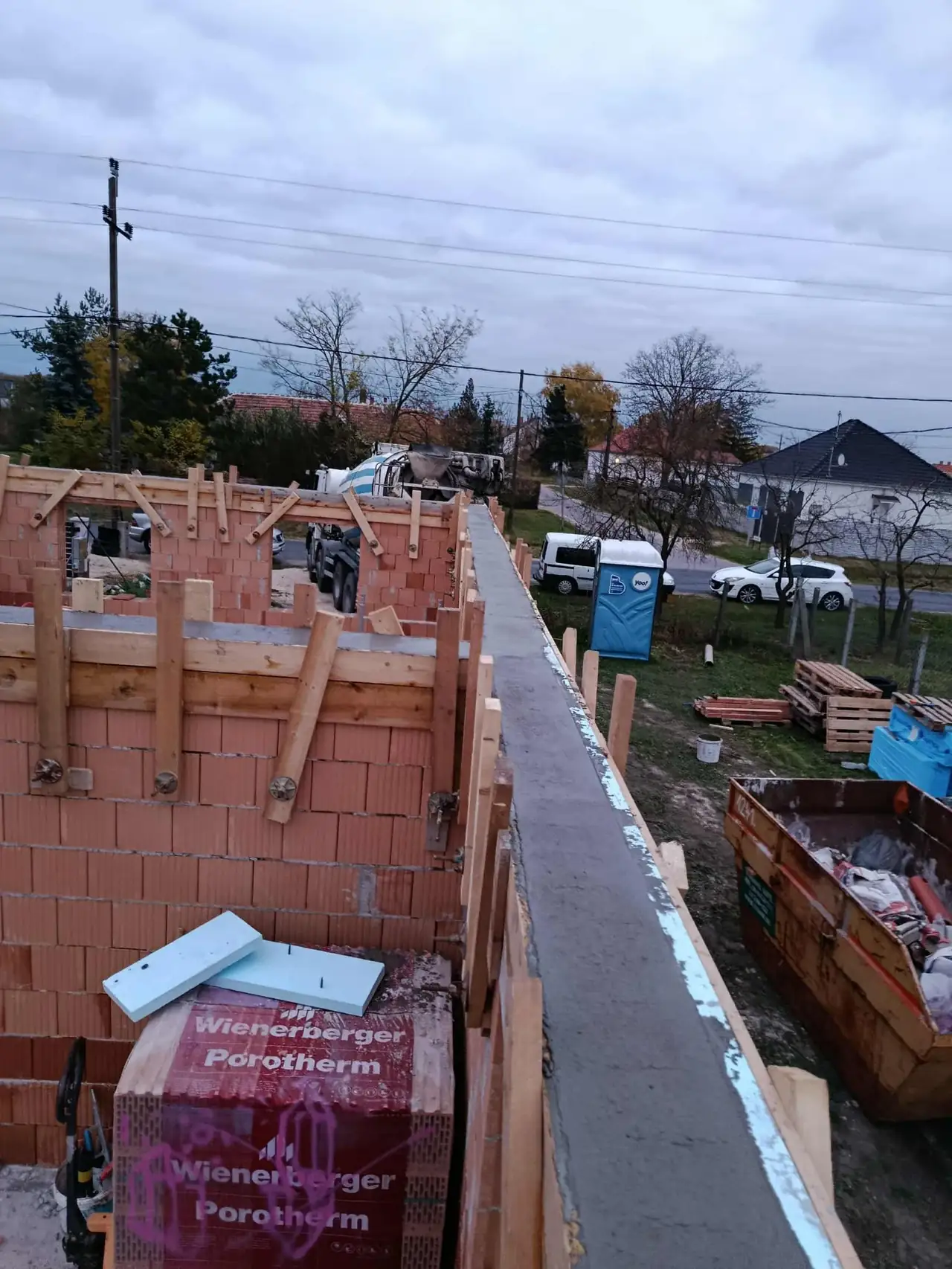 A képen egy építkezés látható Győrben, ahol a Farkas-Konstrukt Kft. generálkivitelezéssel foglalkozik. Az épület falai részben készülnek, téglaanyagokkal és fa állványzattal. A háttérben teherautók és egyéb építőanyagok láthatók, míg a felhős égbolt a munkaterület felett terül el. Az építkezés környezete rendezett, ami a precíz kivitelezést tükrözi.