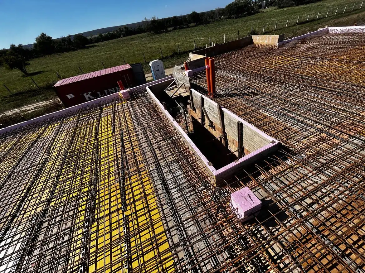 A képen a Farkas-Konstrukt Kft. generálkivitelezési munkálatai láthatók Győrben. A területen egy épület tetőszerkezete látható, amelyen acélgerendák és formázott elemek találhatók. A háttérben zöld fű és fák láthatók, míg a kivitelezéshez szükséges anyagok, például konténerek is megjelennek. A kép jól tükrözi a cég szakmai tevékenységét és a folyamatban lévő munkát.