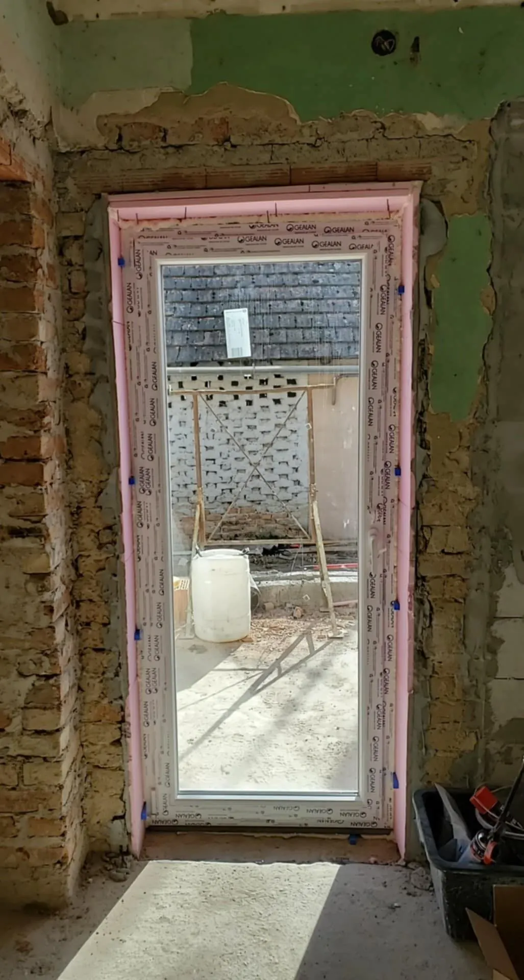 A képen egy építkezés látható, amelyen a Farkas-Konstrukt Kft. generálkivitelezési munkálatai zajlanak Győr városában. A falon egy frissen beszerelt ajtókeret található, körülötte látható a falazás és az építkezés előkészületei. A háttérben építőanyagok és egy állvány látható, ami a munkálatok során használt eszközöket tartalmaz.