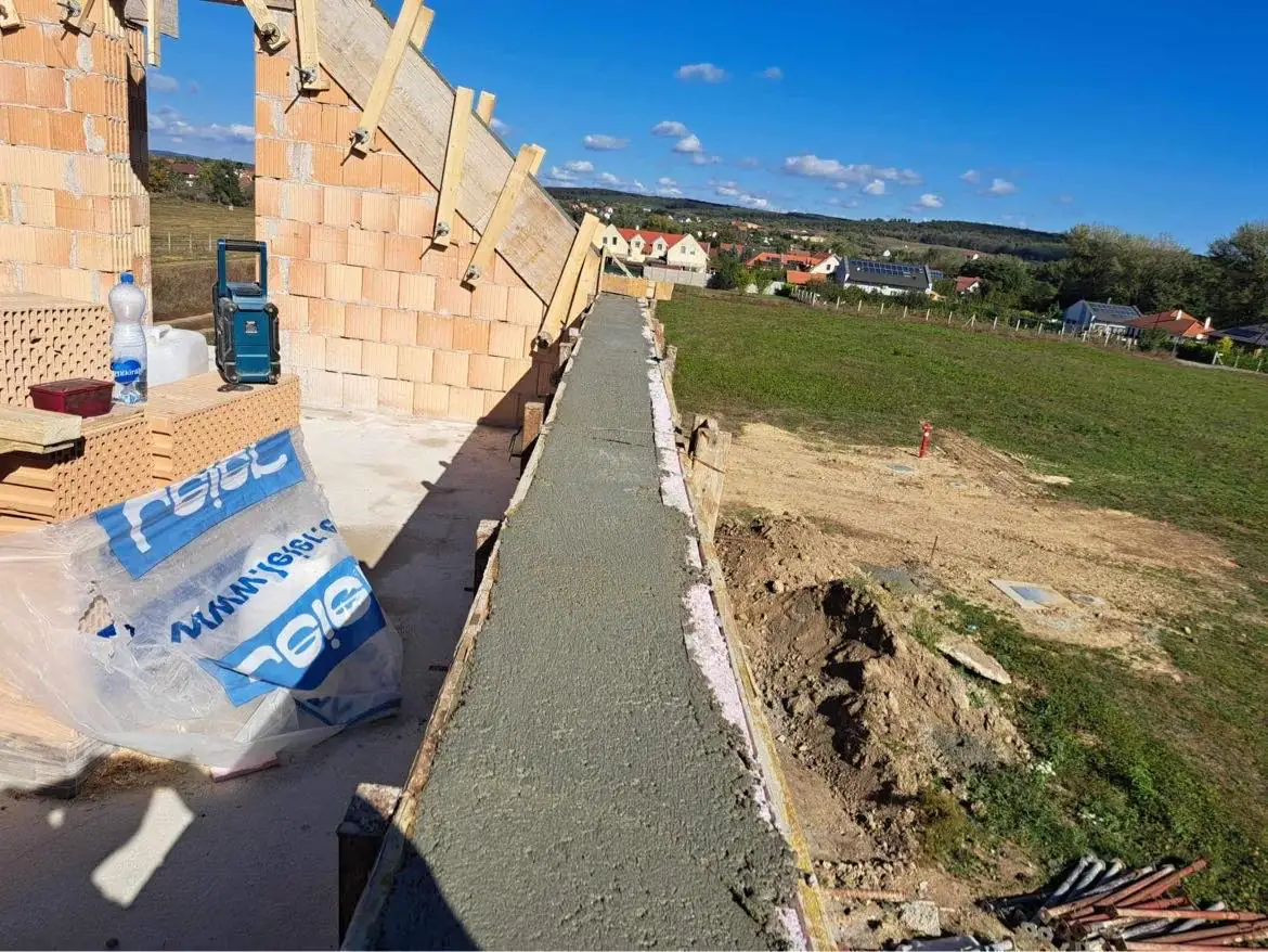 A képen a Farkas-Konstrukt Kft. generálkivitelezés munkálatai láthatók Győr közelében. Az építkezés helyszínén frissen öntött beton van egy hosszú, keskeny járdán, amely a ház tetejére vezet. A háttérben zöld dombok és lakóházak körvonalai látszanak, míg az előtérben építőanyagok, például vízpalack és műanyag zsákok találhatóak.