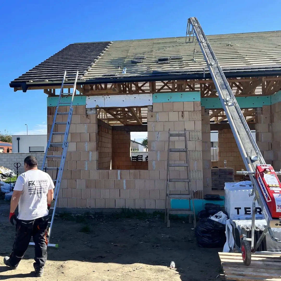 Új tetőfedés folyamatát mutatja be a Farkas-Konstrukt Kft. generálkivitelezési projektje Győrben. A képen látható egy félig elkészült ház, amelynél létrák és tetőfedő eszközök találhatók. A munkás a tetőn dolgozik, miközben a háttérben építőanyagok és egy teherautó látható. Az idő napos, ami kedvez a kivitelezéshez.