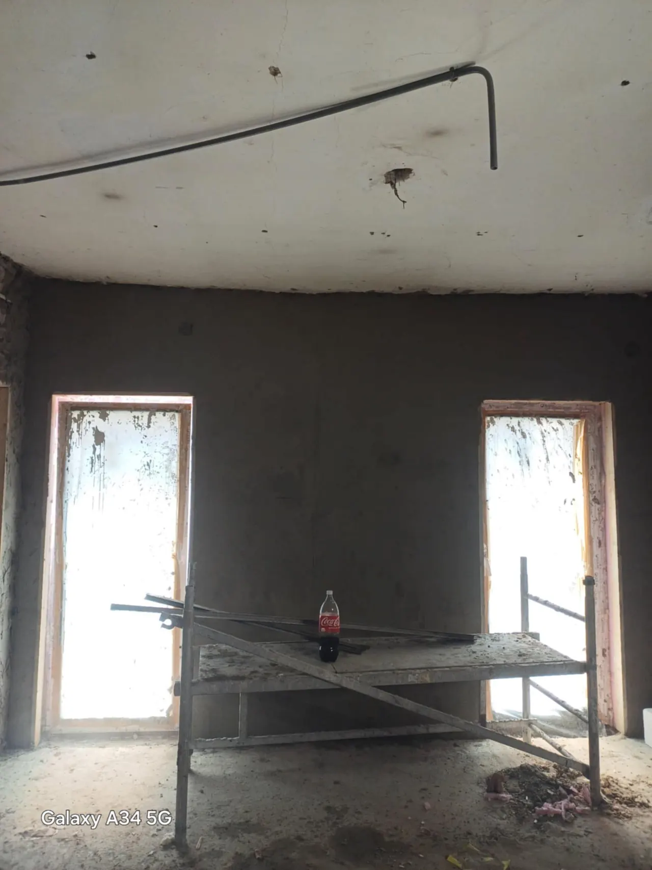 A képen egy családi ház belső terét látjuk, amely még építés alatt áll. A falak simára vakoltak, és két ablak kerete látható, melyek mögött a természetes fény bevilágít. Az építkezéshez használt állványon egy üveg kóla található, ami a munka közben készült pihenőidőt jelzi. A Farkas-Konstrukt Kft. generálkivitelezési szolgáltatásai Győr városában segítik az otthonteremtést.