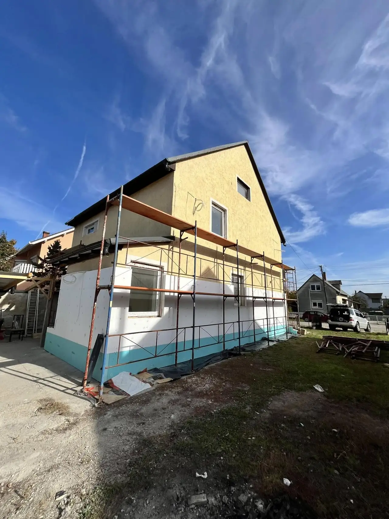 Győr városában, a Farkas-Konstrukt Kft. generálkivitelezési projektjénél egy ház külső homlokzati felújítása zajlik. A képen látható a felújítás alatt álló épület, amelyet állványzat vesz körül. A falak részben készülnek, és a szép kék színű alapozás már látható. Az ég tiszta és kék, ami kedvező időjárást jelez a munkához.