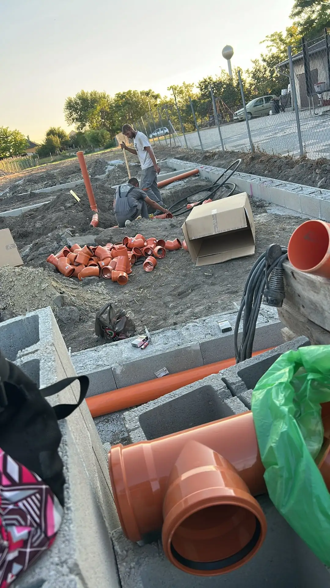 A képen a Farkas-Konstrukt Kft. generálkivitelezési munkálatai láthatók Körmenden. Az építkezés területén munkások dolgoznak, miközben piros csöveket és idomokat helyeznek el a földbe. A háttérben építkezési anyagok, például dobozok és zsákok láthatók, jelezve a folyamatban lévő fejlesztéseket.