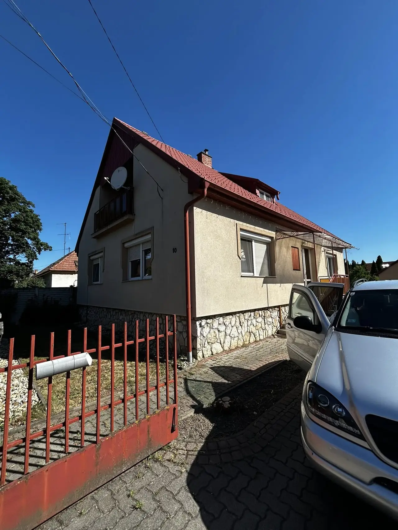 A képen egy győri családi ház látható, amelynek homlokzata világos színű, és kővel burkolt alapja van. A tető piros cserépből készült, és egy antennát is láthatunk rajta. Az udvarban parkoló autó és egy piros kerítés található. A Farkas-Konstrukt Kft. generálkivitelezési szolgáltatásai garantálják az ilyen jellegű építkezések minőségét Győr területén.