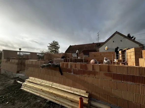 Győri építkezés során a Farkas-Konstrukt Kft. generálkivitelező munkatársai láthatók, amint téglafalakat építenek. A képen a munkások aktívan dolgoznak a falak kialakításán, míg az építkezés körüli terület rendezett faanyaggal van ellátva. A háttérben egy klasszikus magyar ház is látható, ami a helyi építkezések jellegzetességét tükrözi.