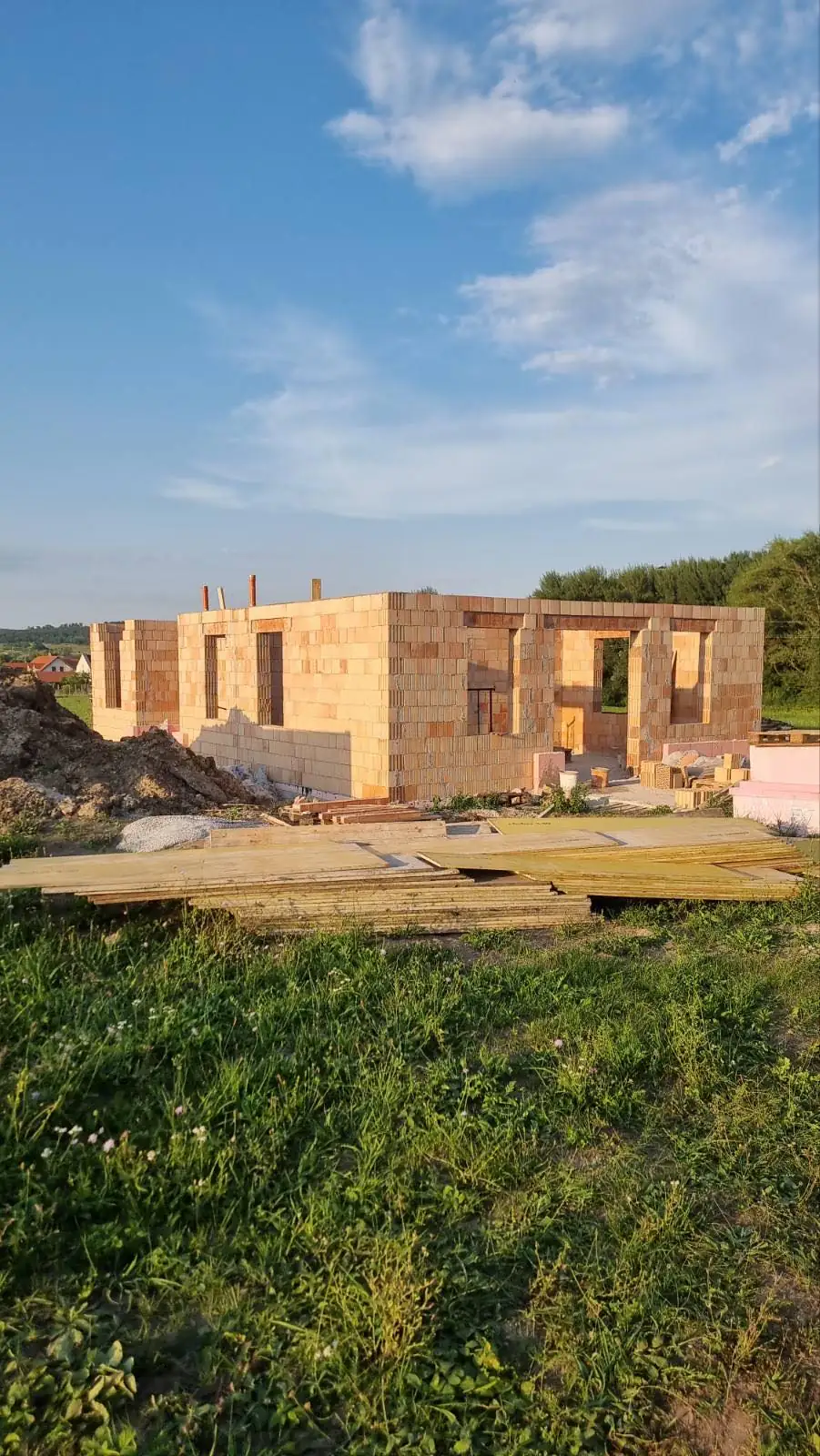 Győr városában a Farkas-Konstrukt Kft. generálkivitelezési szolgáltatása keretében falazás folyik. A képen egy félig kész ház látható, amelynek tégla falai már kialakultak, és a környező területen építkezési anyagok, mint például fából készült deszkák és kövek találhatók. Az építkezés zöldellő tájban helyezkedik el, tiszta kék égbolttal a háttérben.