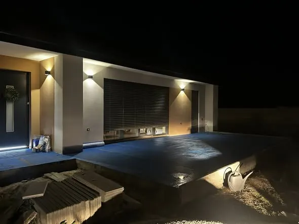 Modern családi ház külső nézete Győrben, amelyet a Farkas-Konstrukt Kft. generálkivitelezett. Az épület előtt szépen megvilágított terasz található, a falak színvilága modern és elegáns. A házhoz tartozó kert rendezett, a terasz mellett egy locsolókanna is látható, ami a gondozott környezetet tükrözi.