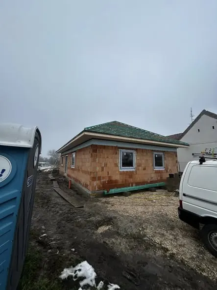 A képen egy épülő ház látható, amelynek homlokzata téglával burkolt. Az épület mögött egy fehér furgon parkol, a bal oldalon pedig egy mobil toalett található. Az idő borongós, és a föld nedves, ami az építkezés fázisára utal. A kép Győr városában készült, ahol a Farkas-Konstrukt Kft. generálkivitelezési munkáit végzi.