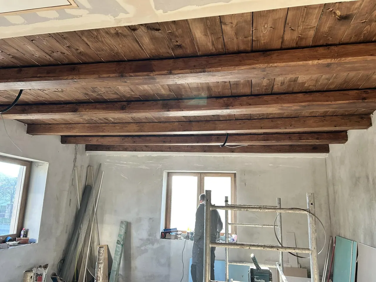 A képen egy Kőszegen található generálkivitelezési projekt belső terét látjuk, ahol fából készült gerendák díszítik a mennyezetet. A falak még festésre várnak, és a munkálatok során állványt is használnak. Az ablakon beáramló fény világítja meg a teret, ahol a Farkas-Konstrukt Kft. munkatársa éppen dolgozik a befejezésen.