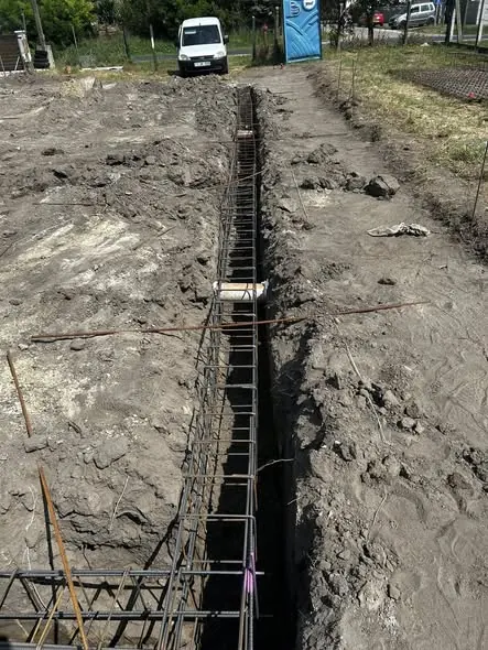 Az image a Fertőd városában található Farkas-Konstrukt Kft. generálkivitelezésének egyik építkezési helyszínét mutatja be. A képen egy mély árok látható, amelyben acélbeton szerkezet készül, a földmunkák és az előkészületek jól láthatók. A háttérben egy fehér furgon parkol, míg a területen építkezési eszközök és anyagok találhatók.
