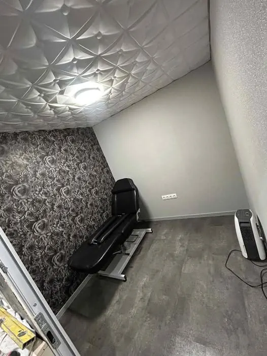 Ez a kép egy modern, jól megtervezett helyiséget mutat be, amely Sárváron található. A falon elegáns, virágmintás tapéta látható, míg a padlón szürke laminált burkolat van. A középen elhelyezkedő fekete kezelőágy tökéletesen illeszkedik a térhez, amely a Farkas-Konstrukt Kft. Generálkivitelezésének egyik projektje. A világítás és a szellőzőberendezés modern megoldásokat tükröz.