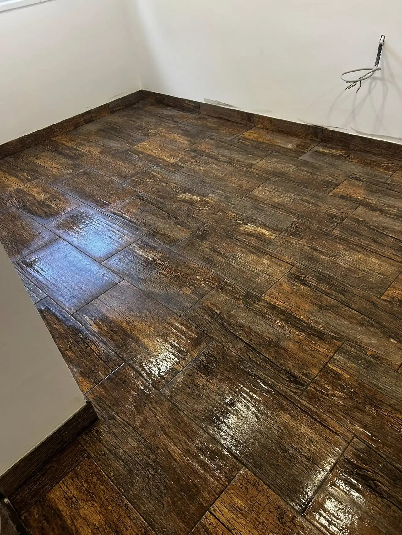 A képen egy frissen lerakott, fényes, barna színű padló látható, amely a modern generálkivitelezés stílusát tükrözi. A padlóburkolat fából készült deszkákra hasonlít, és a világítás szépen kiemeli a felület textúráját. A háttérben egy fehér fal és egy fali WC látható, ami a Tatabánya városában végzett kivitelezési munkák minőségét bizonyítja. A Farkas-Konstrukt Kft. szakértelme nyilvánvaló a részletekben.