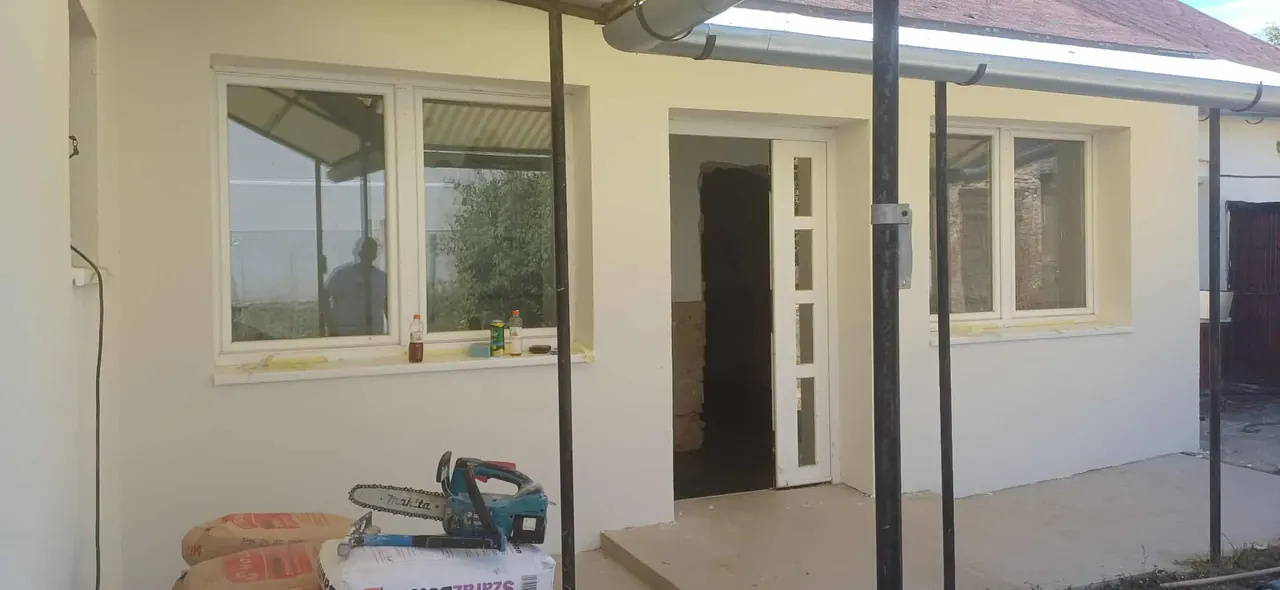 Új építkezési projekt látható a képen, amely a Farkas-Konstrukt Kft. generálkivitelezésében készült Győr városában. A képen egy frissen festett, világos színű ház homlokzata látható, melynek ablakai már be vannak szerelve. Az épület előtt építőanyagok, például cementzsákok és egy láncfűrész található, jelezve a munkálatok folytatását.