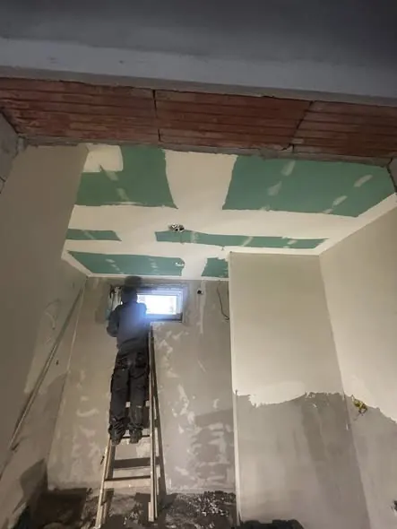 A képen egy munkás látható, amint létrán állva a falon gipszkartonozást végez. A háttérben a mennyezet zöld festékkel jelölt részei láthatók, készülve a végső simításokra. Az építkezés helyszíne Győr városában található, ahol a Farkas-Konstrukt Kft. generálkivitelezési szolgáltatásait végzi. A munkás figyelmesen dolgozik, biztosítva a kivitelezési folyamat zökkenőmentességét.