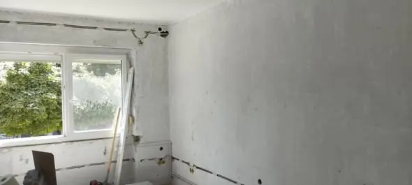 A képen egy felújítás alatt álló belső tér látható, ahol a falak simítatlanok és festésre várnak. Az ablakon keresztül természetes fény árad be, és a kert zöld növényei is látszanak a háttérben. A munkaterületen különféle építőanyagok, például fadarabok és eszközök találhatók, ami a Farkas-Konstrukt Kft. generálkivitelezési tevékenységét tükrözi Komáromban.