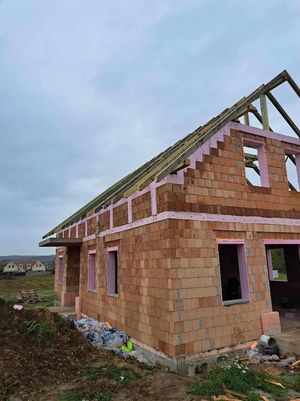 Új építés alatt álló ház látható Győr városában, ahol a Farkas-Konstrukt Kft. generálkivitelezési szolgáltatását végzi. Az épület falai már elkészültek, és a tetőszerkezet is kialakítás alatt áll. A környezet zöldellő, és a háttérben további házak sejlenek fel, amelyek a lakóövezet részei. Az építkezés jól halad, a részletek gondosan kidolgozottak.