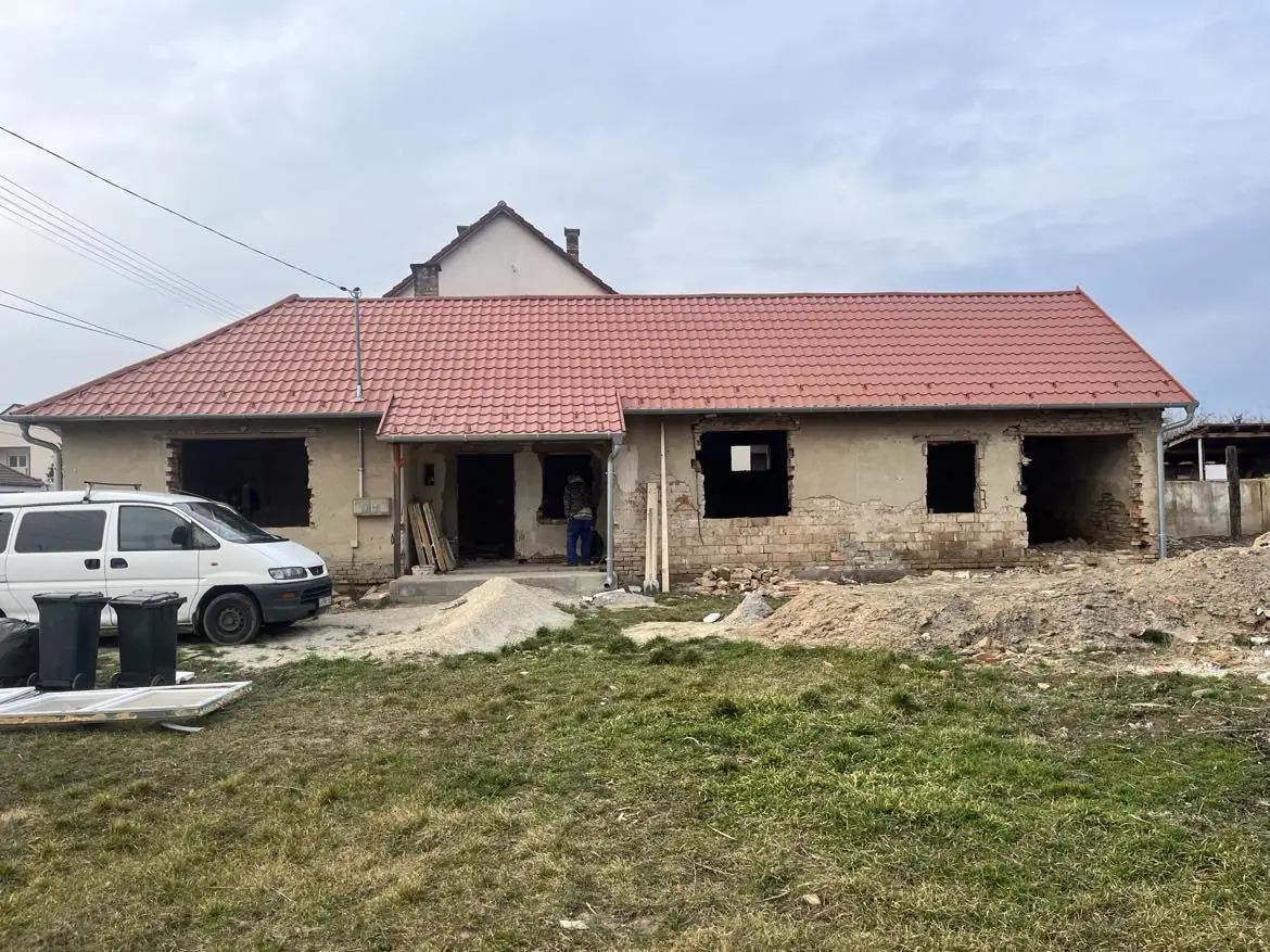 A Farkas-Konstrukt Kft. generálkivitelezési projektje látható Győr városában, ahol egy régi ház felújítása zajlik. Az épület homlokzata már láthatóan készül, az ablaknyílások még üresek, a tető új cseréppel van borítva. A környező területen építőanyagok és munkagépek találhatók, a háttérben pedig egy autó is látható.