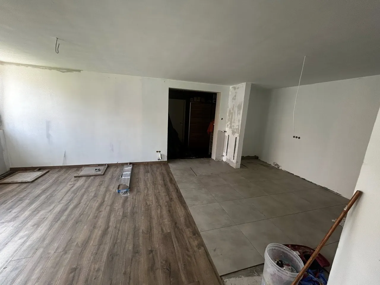 A képen egy félkész lakásbelső látható, ahol a padlóburkolat félig kész, az egyik oldalon fa hatású, míg a másikon csempe található. A falak festésre várnak, és a nyílászárók is be vannak jelölve. A szobában világos színek dominálnak, ami a tágasság érzetét kelti. A Farkas-Konstrukt Kft. generálkivitelezési munkái Szombathelyen folynak.