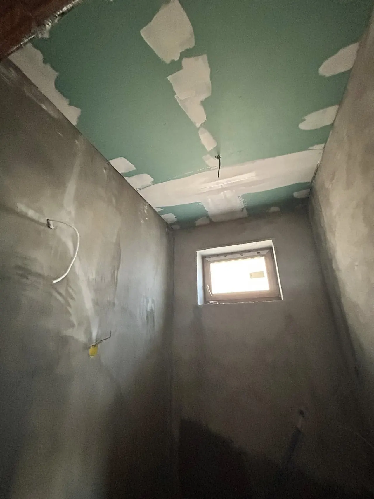 A képen egy építkezés helyszínét látjuk, ahol a falak már meszelve vannak, és a mennyezet frissen előkészített, zöld színű burkolatot kapott. A falakon látható néhány vezeték is, míg a háttérben egy kis ablak található, amelyen keresztül világít be a fény. Ez a munka a Farkas-Konstrukt Kft. generálkivitelezési projektje Győr városában zajlik.