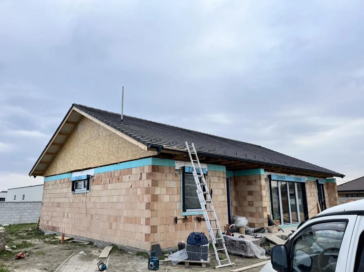 Győr városában, a Farkas-Konstrukt Kft. generálkivitelezés projektjének helyszínén készült a kép, amely egy épülő házat ábrázol. Az épület falai már burkolva vannak, és a tető is megkezdődött. Az építkezés területén különböző szerszámok és anyagok találhatóak, a létra pedig a tetőhöz vezet, jelezve a folyamatban lévő munkát.