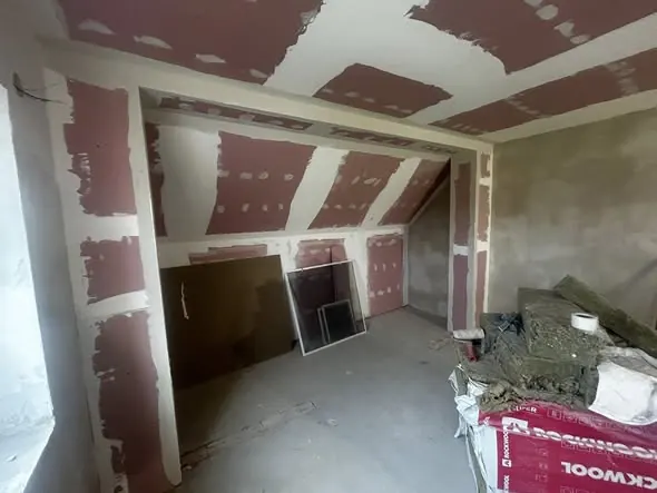 A képen egy generálkivitelezés alatt álló belső tér látható, ahol a falakat és a mennyezetet még javítják. A falakon friss glettelés nyomai láthatók, míg a padlón építőanyagok és szerszámok hevernek. Az építkezés Győr városában zajlik, a Farkas-Konstrukt Kft. vállalat által. A helyiség világos, és a munkálatok előrehaladása jól látható.