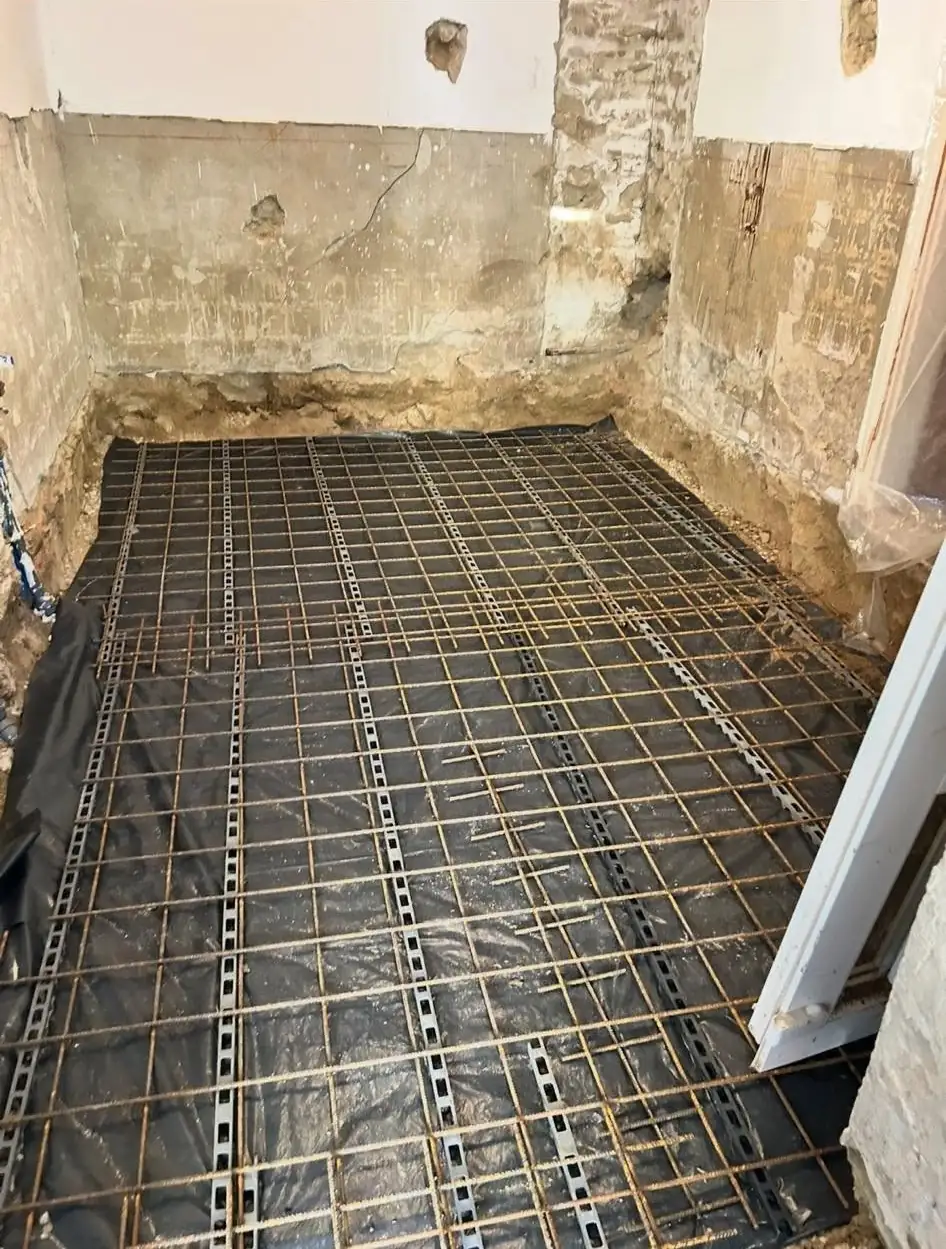 Az image a Farkas-Konstrukt Kft. generálkivitelezési projektjét mutatja be Sárváron. A képen látható egy felújítás alatt álló helyiség, ahol a padló alján fekvő fekete fólia és a ráhelyezett acélrács jól látható. A falakon a régi kő és vakolat nyomai látszanak, ami a munka előrehaladását jelzi.