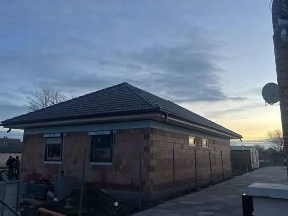 A képen egy győri építkezés látható, ahol a Farkas-Konstrukt Kft. generálkivitelezési munkálatai zajlanak. Az épület már a tetőszerkezetet is magában foglalja, és a falazás előrehaladott állapotban van. A háttérben a naplemente színei látszanak, ami hangulatos atmoszférát teremt az építkezés körül.