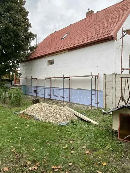 Győr városában, a Farkas-Konstrukt Kft. generálkivitelezési projektjén a ház hátsó oldalán látható a frissen felújított fal, amelyet állványok támasztanak alá. Az építkezés területén kőrakás és fából készült anyagok találhatók, a zöld fű és a fák környezetében. Az épület tetőszerkezete piros cserépből készült, ami modern megjelenést kölcsönöz a háznak.