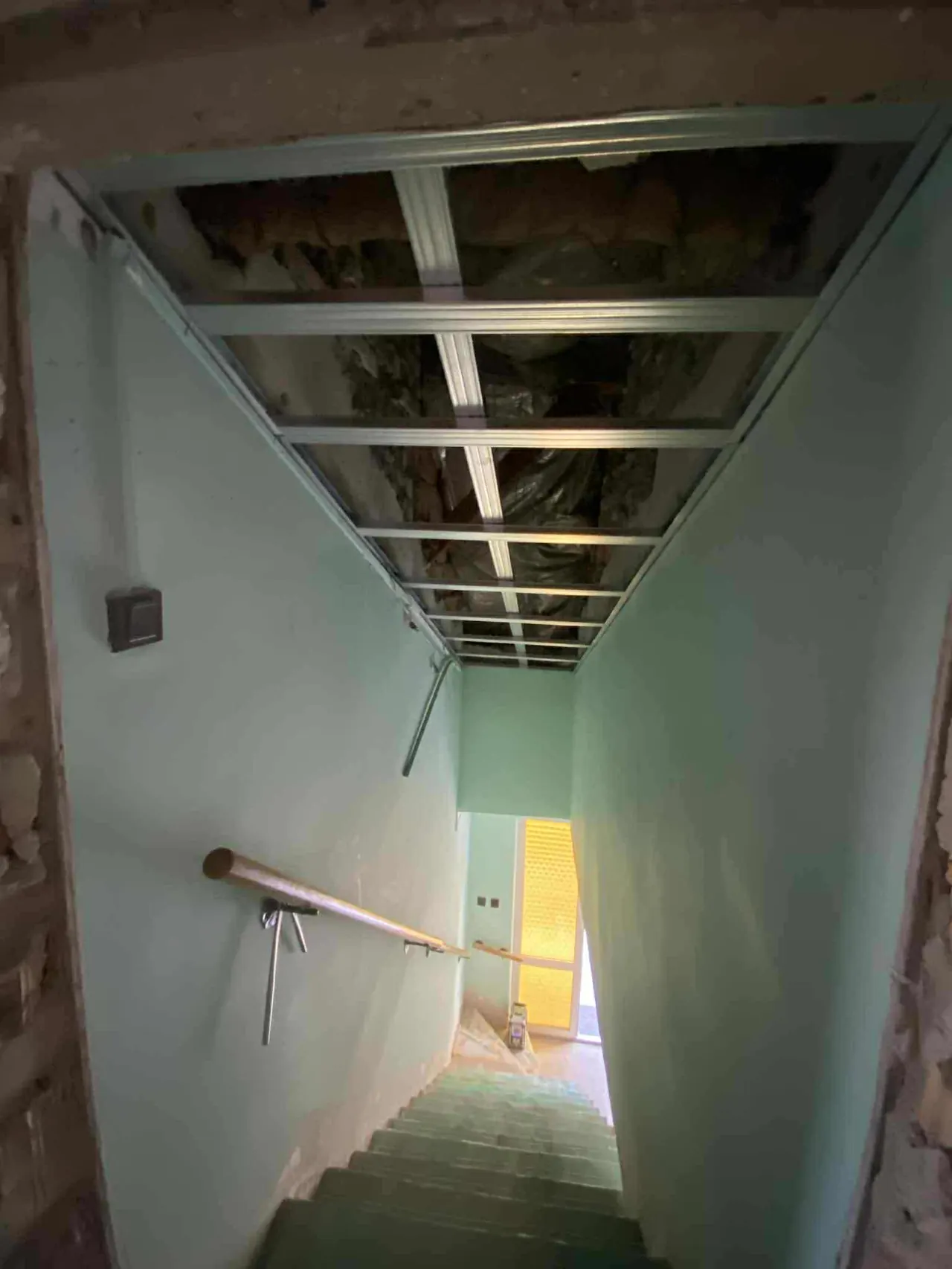 A képen egy lépcsőház belső része látható, amelynek falai világos zöld színűek. A lépcsők fából készültek, és egy fémkorlát segíti a biztonságos közlekedést. A lépcső felé vezető úton természetes fény áramlik be egy nyitott ajtón. A felújítás alatt álló terület Kapuvár városában található, ahol a Farkas-Konstrukt Kft. Generálkivitelezés szolgáltatásait nyújtja.