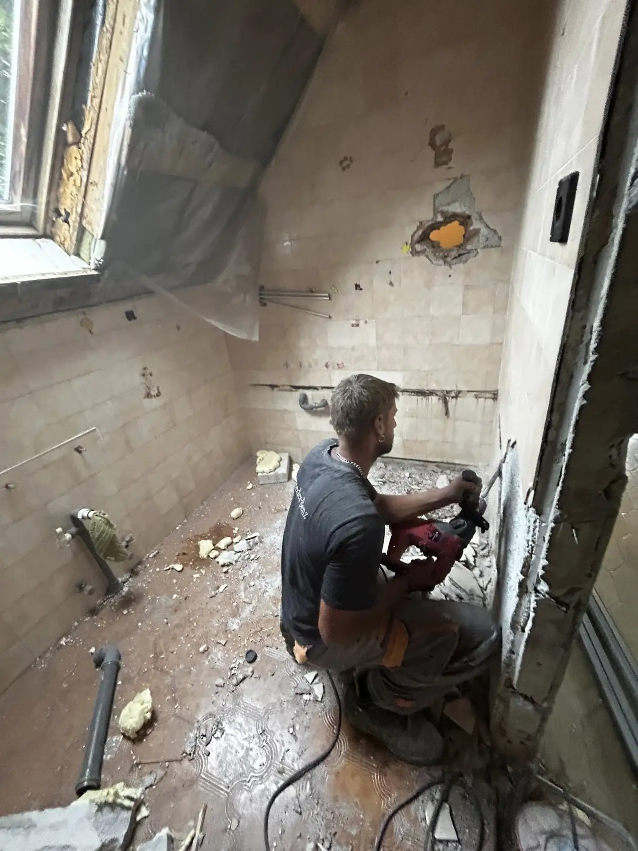 Győr városában, a Farkas-Konstrukt Kft. generálkivitelezés szolgáltatásának keretében egy fürdőszoba felújítási folyamat látható. A képen egy munkás látható, aki éppen a falat készíti elő a felújításhoz, a helyiség falai kopottak és törmelékek borítják a padlót. Az ablakon keresztül beáramló fény a munkafolyamatot világítja meg, hangsúlyozva a renoválás szükségességét.