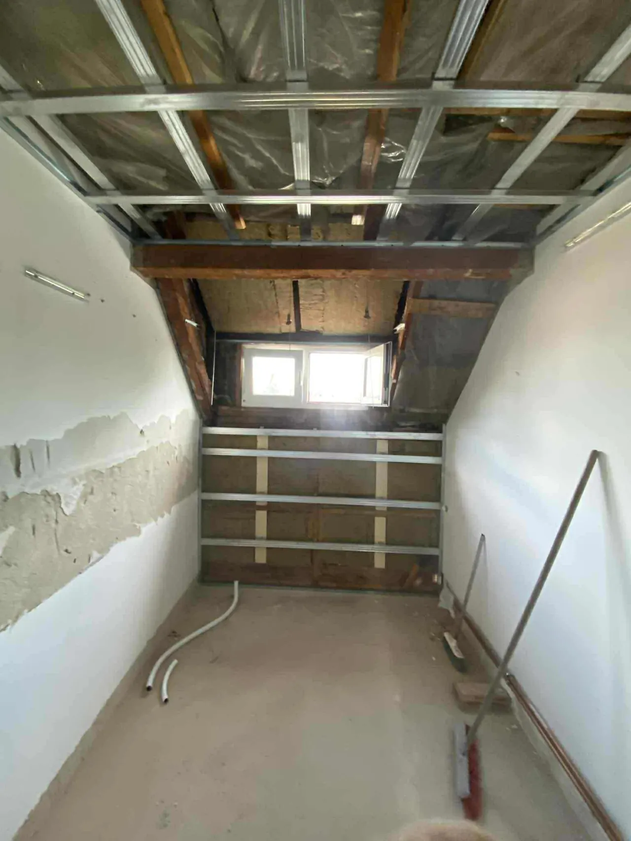 A képen egy tetőtér beépítése látható, ahol a falak és a tetőszerkezet közötti teret alakítják ki. A szobában új ablakok kerülnek beépítésre, és láthatóak a falak burkolásának előkészületei is. A munkaterület rendben van, a padló még nem kész, de a vezetékek már elő vannak készítve. A projekt a Farkas-Konstrukt Kft. generálkivitelezésének része Győr városában.