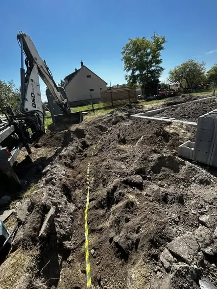 Farkas-Konstrukt Kft. földmunkát végez Győr területén, ahol egy nagy méretű ásó gép látható, amint egy építkezés előkészítése zajlik. A képen a gödörben látható sár és törmelék, valamint a háttérben egy ház áll. A munkaterület rendezett, és a földmunkálatok célja az alapozás elősegítése a generálkivitelezés folyamatában.