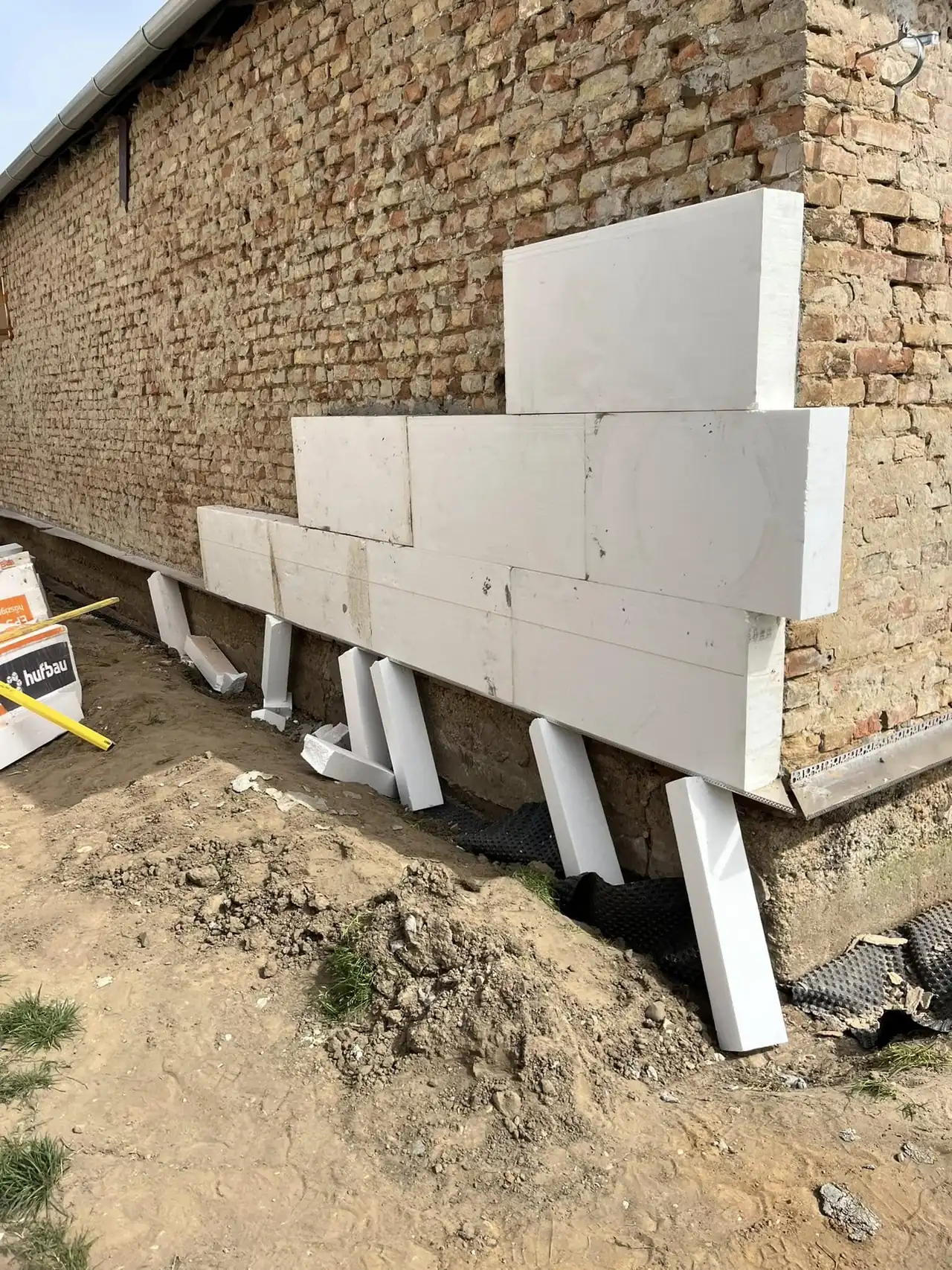 Az image egy építkezés helyszínét mutatja be Győrben, ahol a Farkas-Konstrukt Kft. generálkivitelezési munkálatai zajlanak. A képen látható egy téglafal, amely előtt fehér építőelemek állnak, különböző méretű és formájú darabokból álló struktúrával. A talajon építőanyagok és szerszámok is láthatók, ami az aktív munkafolyamatot jelzi.