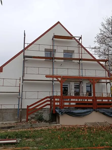 A képen egy részben felújított ház látható, amely Győr városában található. A ház homlokzatát frissen festették, és állványzat borítja, amely a generálkivitelezés munkálatainak része. Az épület előtt egy fa terasz található, amely már kész van, és a kertben némi falevél látható a földön. A háttérben fák és szürke égbolt látható, ami az őszi időszakra utal.