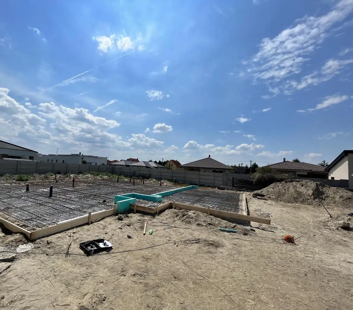 A képen egy építkezési terület látható Győrben, ahol a Farkas-Konstrukt Kft. generálkivitelezési munkálatai zajlanak. Az előkészített földmunkák között látható a betonozásra előkészített alap, amely körül sok építőanyag és szerszám található. Az ég kék, néhány felhővel, ami kellemes időt jelez az építkezéshez.