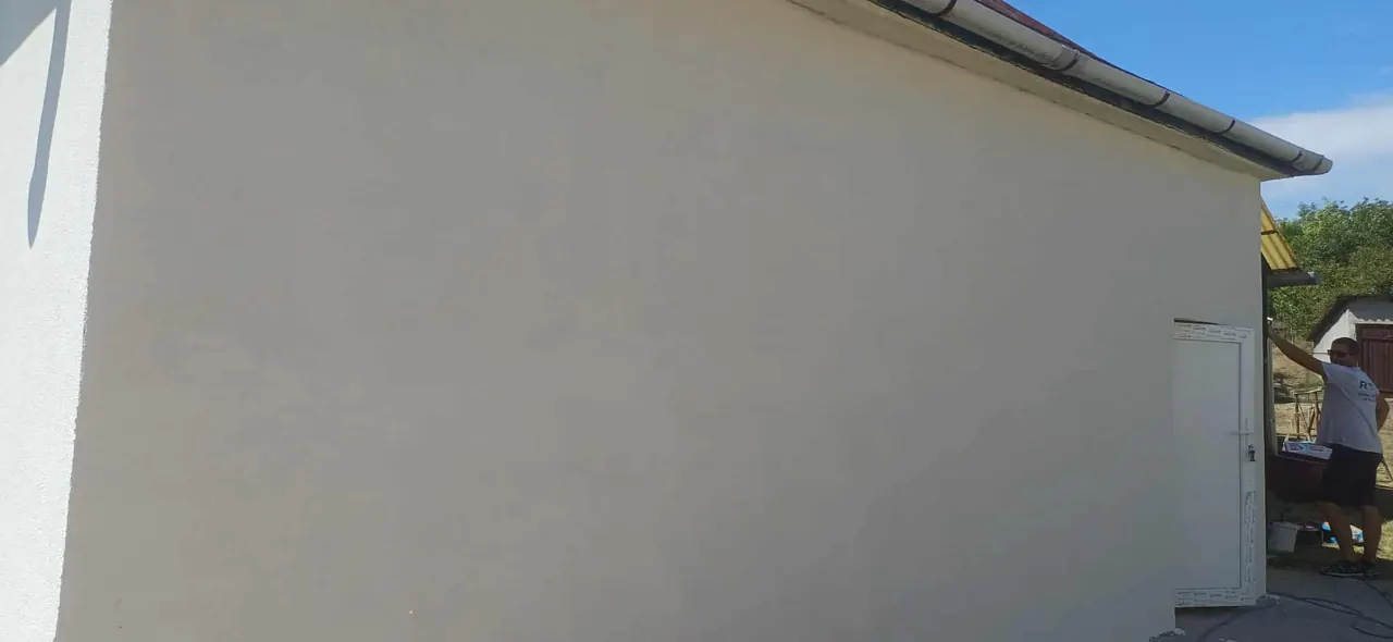 Egy frissen vakolt fal látható, amelynek sima, világos felülete tűnik ki. A háttérben egy felhőtlen kék égbolt látható, ami élénk kontrasztot ad a fal színével. Az építkezésen dolgozó szakember éppen a fal utolsó simításait végzi, ami a Farkas-Konstrukt Kft. Generálkivitelezés szolgáltatásának minőségét tükrözi Mosonmagyaróváron.