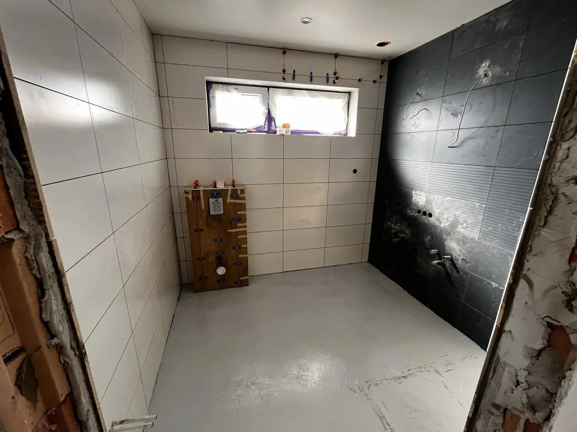 A Farkas-Konstrukt Kft. által végzett fürdőszoba felújítás folyamatának képe, amely Győr városában készült. A képen látható a frissen burkolt fal, a csempe és a padló, valamint az előkészített vízvezetékek. A munkaterület rendezett, a világos és sötét csempe kontrasztja modern megjelenést kölcsönöz a fürdőszobának, amely a generálkivitelezés során készült.