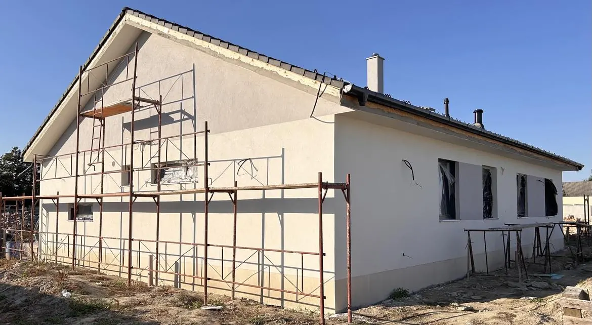 A képen egy Bük városában folyó generálkivitelezési munka látható. Az épület oldalán frissen felhordott vakolat és ablaknyílások láthatók, melyek körül állványzat van felállítva. A munkálatok során a Farkas-Konstrukt Kft. szakemberei gondoskodnak a kivitelezés precizitásáról. A háttérben a tiszta kék égbolt és a környező terület is jól látható.