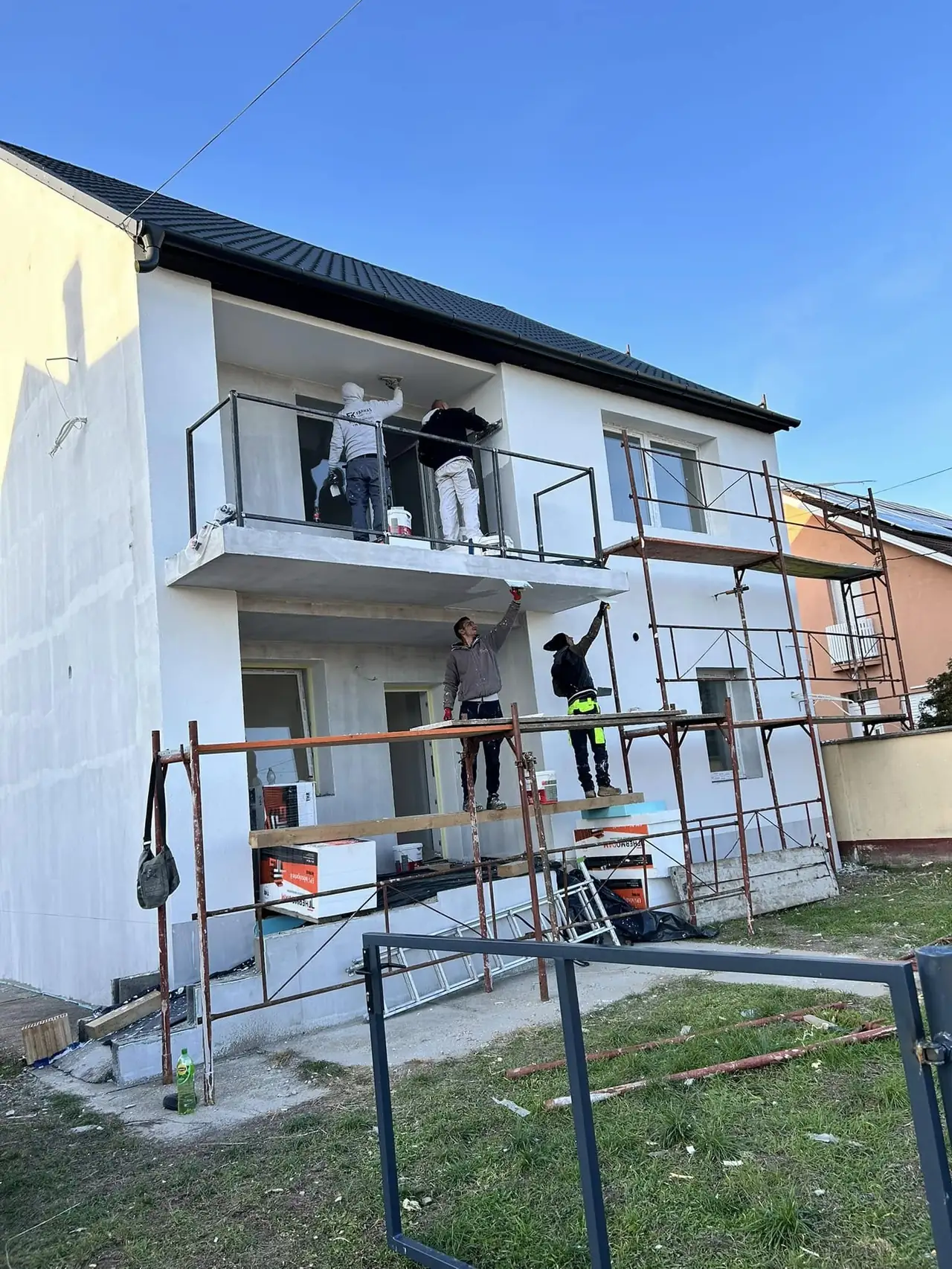 Győr városában a Farkas-Konstrukt Kft. generálkivitelezési projektje zajlik, ahol munkások egy új épület homlokzatát festik. Az épület előtt állványzat található, amelyen több szakember dolgozik, míg egy másik munkás a földön segít. A világos égbolt és a napsütés ideális körülményeket biztosít a munkához, amit a cég precíz kivitelezése jellemez.