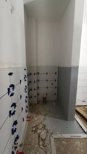 A képen egy építkezés látható, ahol a falakat csempézik. A csempék már elhelyezésre kerültek, és a fal alsó részén szürke színű burkolat látható. Az építkezés helyszíne Fertőd, ahol a Farkas-Konstrukt Kft. generálkivitelezési munkálatokat végez. Az építkezés során a precíz munkavégzés és a szakszerű kivitelezés kiemelten fontos.