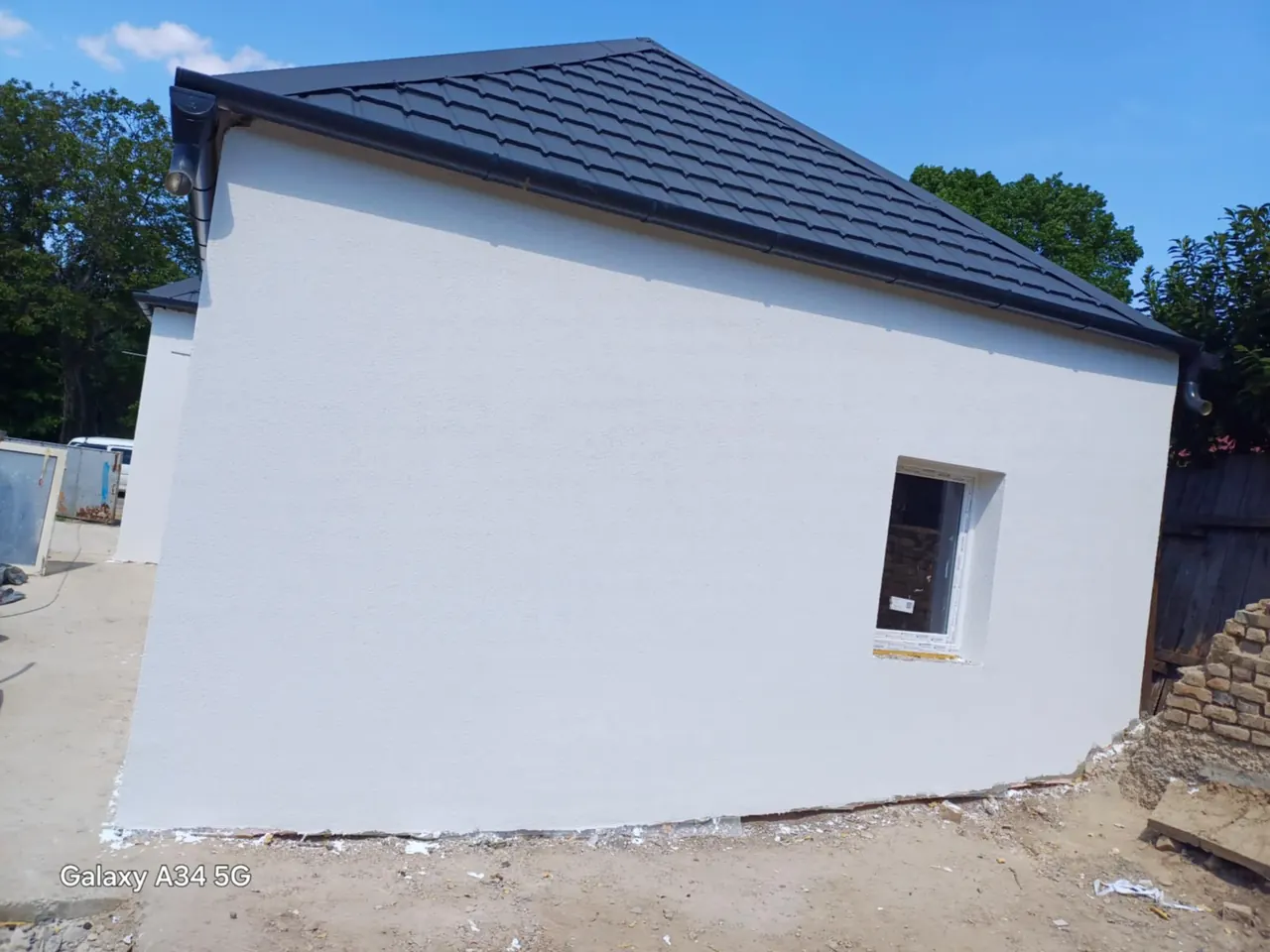 A képen egy frissen épített ház látható, amelynek sima, fehér falai és sötét cseréptetője van. A ház oldalán egy ablak található, amely még nem lett befejezve. A környezetben építési törmelék és föld látható, ami a generálkivitelezési munkálatok folyamatát tükrözi. A helyszín Győr városában található, ahol a Farkas-Konstrukt Kft. végzi a kivitelezést.