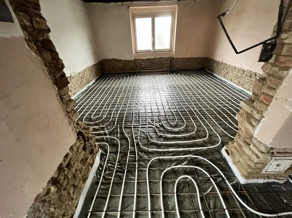 A képen egy Győr városában folyó generálkivitelezési projekt látható, ahol a hőszigetelés munkálatai zajlanak. A szobában a padló alá telepített fűtőcsövek láthatók, amelyek precízen elhelyezve futnak a padlófelületen. A falak részben látszanak, a téglák és a vakolat kombinációjával. A világítás természetes, a szoba ablakán át beszűrődő fény világítja meg a munkaterületet.