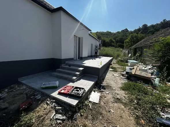 A képen a Farkas-Konstrukt Kft. generálkivitelezési projektje látható Győr városában. A felvételen egy frissen épített terasz látható, amely lépcsőkkel és egy simított felülettel rendelkezik. A háttérben zöld növényzet és épületek látszanak, míg a terasz körül építkezési hulladék és szerszámok találhatók, ami a munkálatok folyamatát tükrözi.