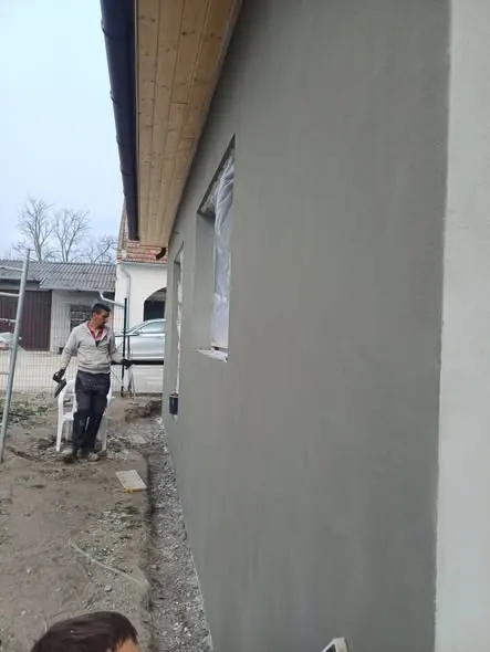 A képen a Farkas-Konstrukt Kft. generálkivitelezési munkálatai láthatók Győrben. Az épület falának simítása zajlik, miközben a munkás figyelmesen dolgozik. A háttérben egy másik épület és kerítés is megfigyelhető, ami a helyszín építkezési jellegét hangsúlyozza. A terület rendezett, és a munka előrehaladása jól látható.