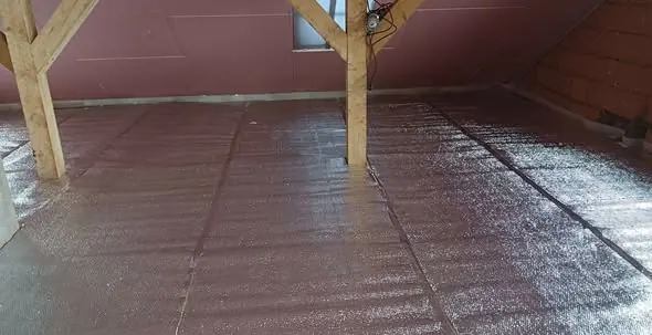 A képen egy Tata városában végzett generálkivitelezés látható, amely során egy tetőtér szigetelése készül el. A padló sima, fényes felületű, ami a szigetelőanyag alkalmazására utal. A fa tartószerkezetek és a falak részletei is jól láthatók, amelyek a munka precizitását tükrözik.