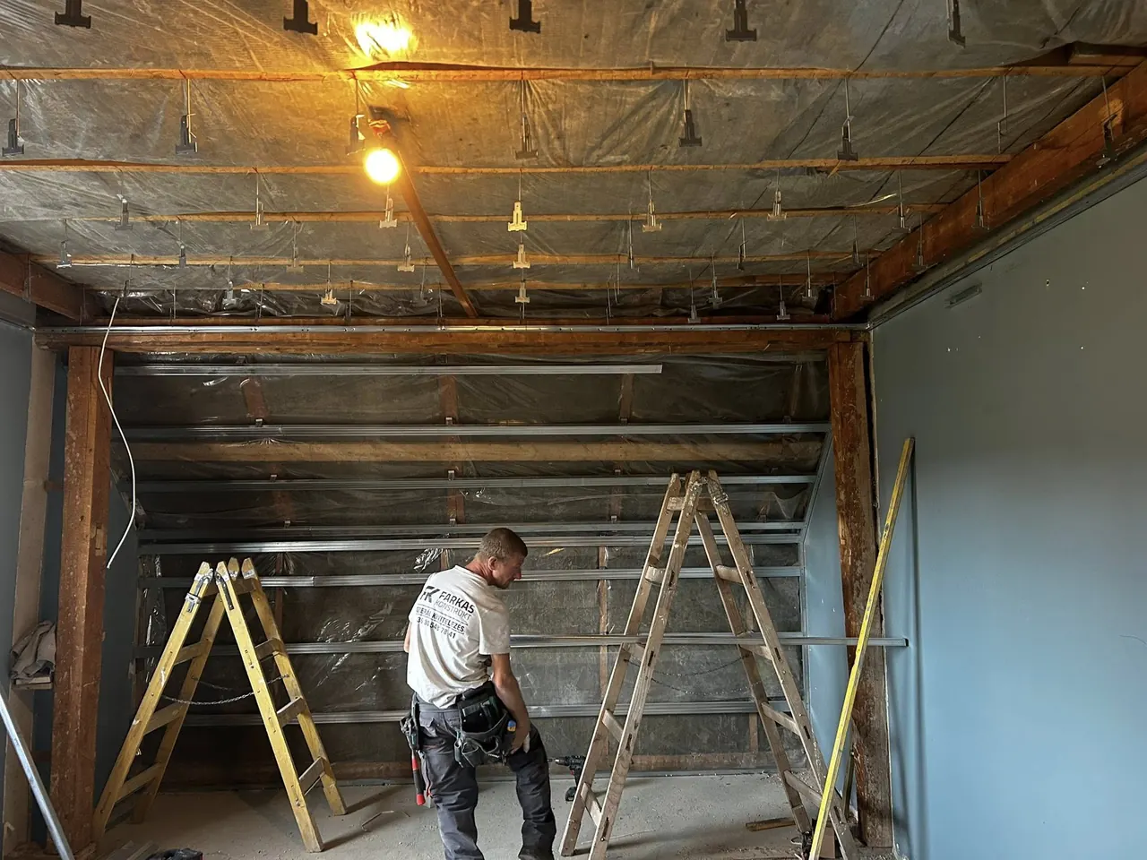 A képen látható egy építkezési terület, ahol a Farkas-Konstrukt Kft. generálkivitelezési munkái zajlanak Győr városában. A háttérben állványok láthatók, míg egy szakember dolgozik az új családi ház tervezésén. A mennyezeten lámpák és szigetelőanyagok találhatók, ami a folyamatban lévő építkezés állapotát tükrözi.