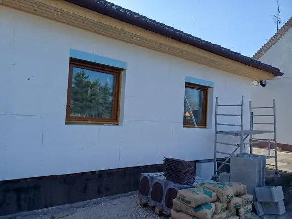 Új építésű, jól megvilágított ház külső fala látható, amelyen kék keretes ablakok találhatók. Az építkezés területén különböző építőanyagok, például csempe és betonblokk halmozódik. A háttérben állvány található, amely a munkálatok előrehaladását segíti. A Farkas-Konstrukt Kft. által végzett generálkivitelezés Győr városában zajlik, ahol a modern építkezési megoldások érvényesülnek.