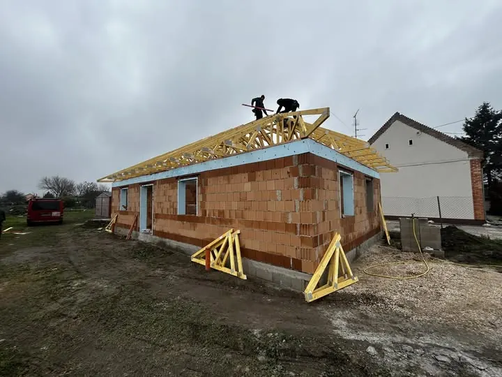 A képen egy épülő ház látható, amelyen két munkás dolgozik a tetőszerkezet kialakításán. A falak téglából épültek, és a tető gerendái már a helyükre kerültek. A környék szürke időjárással borított, a háttérben egy másik épület is látszik. A Farkas-Konstrukt Kft. Generálkivitelezés tevékenysége Győr városában zajlik, ami a fejlődő lakóházak minőségi kivitelezését jelzi.